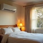 A white wall-mounted split system air conditioner in a softly lit bedroom at dusk, with a made bed, a bedside lamp and gum trees outside the window