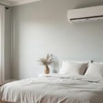 A plain white wall-mounted split system indoor unit above a made bed in a light grey bedroom, with a gum tree visible through the window