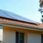 Rooftop solar panels on a terracotta tile roof above a white split system air conditioner outdoor unit on a rendered wall, with a gum tree behind
