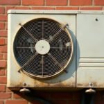 A weathered off-white air conditioner outdoor unit with rust along its base, mounted on a red brick wall